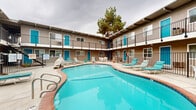 Located at 911 Pennsylvania Place in Davis, California, this mid-century style multi-unit property is centered around a striking, freeform outdoor swimming pool that serves as the focal point of the complex. The pool's curved, kidney-inspired shape is bordered by a brick-toned coping and a generous concrete deck outfitted with teal lounge chairs and patio seating, creating a welcoming communal outdoor retreat.

The two-story building wraps around three sides of the courtyard in a classic U-shaped configuration, offering residents direct visual access to the pool area. Each floor features individual unit entrances with teal-painted doors — a consistent design accent that lends the property a cohesive, retro aesthetic. Upper-floor units open onto a shared balcony walkway lined with black iron railings, while ground-floor units access the pool deck directly. The exterior is finished in a warm taupe stucco with wood beam detailing along the roofline, consistent with mid-century architectural styling.

The open courtyard benefits from mature trees that provide natural shade and a sense of established landscaping. A black iron perimeter fence encloses the pool area for safety. The property is situated within the city of Davis, CA, close to the University of California, Davis campus, making it a well-positioned residential complex in a vibrant college community.