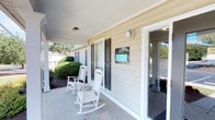 The Park Apartments Clubhouse exterior presents a warm and inviting first impression, designed with classic residential charm in mind. The single-story structure features light-colored lap siding complemented by dark shutters, giving the building a timeless, neighborhood-friendly aesthetic. A covered front porch stretches along the entrance, offering a comfortable and shaded gathering space for residents. Two classic white rocking chairs sit on the smooth concrete porch, inviting visitors to relax and take in the surroundings before stepping inside. The building is framed by neatly manicured shrubs and mature trees, lending a sense of greenery and care to the overall environment. A prominent signage display for the Park Apartments is mounted near the main entrance, clearly identifying the clubhouse as the community's central hub. The entry features glass-paneled doors that allow natural light to filter into the interior while maintaining an open and accessible feel. The overall ambiance is one of casual comfort and community-oriented design, reflective of a well-maintained residential property. The exterior's approachable architecture and thoughtful landscaping create an environment that feels both functional and welcoming for all residents and guests.