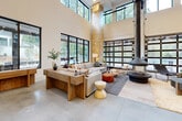 This thoughtfully designed communal space blends industrial and natural elements to create an inviting, multi-functional environment. The centerpiece of the main lounge is a dramatic double-height ceiling that amplifies the sense of openness, anchored by a striking suspended cylindrical fireplace. Floor-to-ceiling black-framed windows and large glass garage-style doors flood the interior with natural light while framing views of the surrounding greenery. Polished concrete floors run throughout, complementing the warm tones of natural knotty pine wood columns, built-in shelving units, and ceiling panels that add organic texture across the space. The lounge area is furnished with generous sectional sofas, solid wood coffee tables, and layered rugs, creating distinct zones for relaxation and conversation. A full bar with a sleek white countertop, navy cabinetry, wall-mounted television, and swivel bar stools serves as a central social hub. An adjacent game area features a billiards table alongside wall-mounted guitars, adding a recreational dimension. A wood-and-steel staircase with a slatted pine partition leads to an upper mezzanine level. Cozy nook seating near oversized windows offers views of an outdoor patio. The overall ambiance is warm, modern, and community-focused, balancing rugged materials with refined finishes.