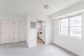 This single-floor unit is laid out with a practical, straightforward floor plan visible across several interconnected rooms. The living area is generously proportioned, finished with neutral gray carpet and cream-toned walls that create a clean, understated atmosphere. A double closet and an additional door provide convenient storage and access directly from this space. Natural light enters through gridded double-hung windows, lending the room a bright, open feel.

The separate kitchen is compact yet fully functional, featuring light blush-toned cabinetry, white countertops, and tile flooring in warm earth tones. It is equipped with an electric range, a dishwasher, a refrigerator, and a sink positioned beneath a window. The layout makes efficient use of the available space.

The bedroom areas are carpeted and well-lit, each with windows that invite natural light. One bedroom benefits from corner windows on two walls, providing an especially airy quality. Closet access is available within the sleeping areas.

The bathroom features a classic black-and-white tile design, with white square wall tiles framed by a bold black border, giving it a timeless, retro aesthetic. An electrical panel is accessible within the unit. Overall, the space presents a neutral, move-in-ready environment with consistent finishes throughout.