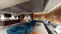 This single-floor amenity lounge is thoughtfully designed with a contemporary aesthetic that blends warm natural materials with modern furnishings. The space is organized into several distinct functional zones that flow seamlessly into one another.

The central lounge area is anchored by a large, curved sectional sofa in charcoal grey, accented with teal and white throw pillows. The sofa wraps around a textured wood-paneled feature wall, which is backlit to create a warm, ambient glow. A sleek arc floor lamp and round wooden coffee table complete this gathering area.

A secondary seating vignette features plush, tufted teal velvet barrel chairs paired with low side tables, while cognac leather accent chairs occupy a quieter reading nook nearby. A curated gallery wall of black-and-white prints adds an artistic focal point to this section.

The bar and kitchenette area includes dark wood cabinetry, a white countertop island with bar seating and pendant lighting overhead, a wall-mounted TV, and a marble tile backsplash with undermount sink. The kitchen counter is accented with decorative brass accessories.

Throughout, grey carpeted flooring, exposed ceiling infrastructure, recessed lighting, and track lighting contribute to the space's modern, urban character.