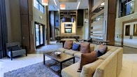 This single-floor space presents a striking, contemporary lobby lounge defined by its dramatic double-height ceilings and refined architectural detailing. The main seating area centers around plush, neutral-toned sofas accented with an array of richly colored throw pillows in deep burgundy, navy, and brown, arranged around a solid wood and metal coffee table set atop a textured area rug. A built-in feature wall serves as the room's focal point, housing an illuminated display niche with open shelving and an integrated media screen, framed by dark, moody cabinetry that adds depth and contrast. Pendant lighting — including a bold cylindrical drum fixture and sleek linear ceiling lights — provides layered illumination throughout the space. Floor-to-ceiling glass entry doors along the perimeter flood the interior with natural light while offering views to the exterior. In the background, a counter-height surface suggests a social or service area, adding to the space's versatile, hospitality-inspired functionality. The overall ambiance is modern and upscale, blending warm wood tones with dark finishes to achieve a polished yet inviting environment.