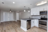 Located at 825 Powder Springs St in Marietta, Georgia, this single-floor unit offers a clean, modern aesthetic with consistent warm-toned hardwood-style flooring running throughout every room. The open-concept layout connects the kitchen and living area seamlessly, creating a bright and functional living environment. The kitchen is well-appointed with crisp white shaker-style cabinetry, dark countertops, a marble-style tile backsplash, and a full suite of stainless steel appliances including a refrigerator, electric range, over-the-range microwave, and a peninsula with pendant lighting that defines the space. The living area is generously proportioned, featuring a stylish ceiling fan with an integrated light fixture and neutral gray walls that lend a calm, versatile backdrop. The bedroom includes a ceiling fan, a dedicated closet, and direct access to additional spaces down the hallway. A secondary room offers flexible use as a bedroom or office, with clean walls and ample floor space. The bathroom is finished with a white vanity, dark countertop, framed mirror with lighted sconces, and a tub-shower combo with marble-style tile surround. Throughout the unit, white baseboards and neutral tones create a cohesive, move-in-ready environment well-suited for comfortable everyday living.