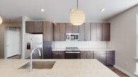 Located at 539 Foster St in Durham, NC, this single-floor unit showcases a cohesive modern design with warm wood-look plank flooring running throughout the space. The open-concept kitchen serves as a central highlight, featuring floor-to-ceiling dark espresso cabinetry, a white subway tile backsplash, and a generous island topped with striking veined quartz countertops. A sculptural globe pendant light anchors the space above the island, while stainless steel appliances—including a French door refrigerator, range, and microwave—complete the contemporary aesthetic.

The living area is bright and open, benefiting from multiple windows fitted with horizontal blinds that allow natural light to filter in while framing views of the surrounding neighborhood. The neutral gray walls and clean white trim create a versatile backdrop throughout.

Two bathrooms serve the unit, each finished with dark wood-toned floating vanities and white countertops. The primary bathroom features a double-sink vanity with an expansive mirror and connects directly to a bedroom. A secondary bathroom includes a tall storage tower for added functionality. The hallways connect rooms efficiently, reinforcing the layout's practical and well-organized flow.