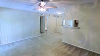 This single-story space features an open-concept layout that connects the main living area to the kitchen through a pass-through counter with a mirrored backsplash, allowing natural sightlines across the space. The living area is carpeted in neutral tones, complementing the light-colored walls that create a bright, airy atmosphere. A ceiling fan with a built-in light fixture is centrally positioned overhead, providing both ventilation and ambient lighting. The pass-through counter, finished with a dark countertop, offers a convenient transition between the kitchen and living areas, with cabinetry visible above. HVAC vents are integrated into the walls, ensuring efficient climate control throughout. A doorway visible in the background leads to an additional room, likely a bathroom or hallway, expanding the overall functionality of the space. White baseboards frame the carpeted floors, adding a clean, finished look. The overall design is practical and straightforward, with a neutral palette that provides a versatile backdrop for various furnishing styles.