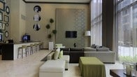 This refined single-floor space exudes a polished, contemporary aesthetic defined by thoughtful design choices and high-end finishes throughout. The main lounge area serves as the focal point, featuring a generous seating arrangement with a modular dark sofa accented by lime green and patterned throw pillows, complemented by a white leather ottoman and a draped green accent table. A large decorative wall panel with an intricate damask-style motif anchors the space, serving as a sophisticated backdrop for the wall-mounted flat-screen television. Soaring ceilings and floor-to-ceiling sheer curtains allow natural light to flood the interior, creating an airy and luminous ambiance. Tall, sculptural indoor plants introduce organic texture and a touch of greenery to the neutral color palette of warm greys, taupe, and olive tones. A set of circular black-and-white classical art prints adorns one wall, adding a cultural and artistic dimension to the space. Adjacent to the lounge, a sleek bar counter with green bar stools offers a dedicated casual seating and refreshment area. The light-toned flooring ties the entire space together, enhancing its sense of openness and cohesion.