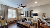 This thoughtfully designed single-floor residence offers a cohesive blend of contemporary style and warm, eclectic touches across its well-appointed rooms. The living area serves as the heart of the space, anchored by a tufted gray sofa, a rustic wood coffee table, and a distinctive gallery wall showcasing eight framed architectural sketches. A ceiling fan with a dark wood finish adds both function and character overhead, while a large window dressed in neutral curtains fills the room with natural light. Rich hardwood-style flooring runs throughout the main living areas, lending warmth and continuity. The kitchen features white shaker-style cabinetry, dark granite countertops, a mosaic tile backsplash, and stainless steel appliances, creating a practical yet polished cooking space. The primary bedroom offers a serene retreat with a metal-framed bed, a substantial dark wood dresser topped with a decorative mirror, and floor-to-ceiling curtains that lend an air of privacy. A second bedroom exudes character with a curated gallery wall arrangement and coordinated dark wood nightstands. The bathroom is finished with white cabinetry, dark granite counters, a large mirror, and a shower-tub combination with clean tile surround, completing this inviting and well-considered home.