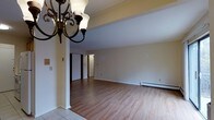 This single-floor, 1-bedroom, 1-bathroom residence offers a straightforward and functional layout designed to maximize everyday comfort. The main living area is open and airy, featuring warm hardwood-style flooring that extends throughout the space, creating a cohesive and inviting atmosphere. A decorative wrought-iron chandelier with classic globe-style shades serves as a focal point near the entryway, adding a touch of traditional charm to the otherwise clean, neutral interior. The walls are finished in crisp white tones, contributing to a bright and open feel. A structural column subtly defines the transition between the entry and the main living area. Baseboard heating units line the perimeter, ensuring efficient climate control throughout the colder months. The entryway features light-toned tile flooring, providing a practical transition zone. Doorways visible toward the rear of the space lead to additional rooms, including the bedroom and bathroom, offering a sensible and accessible flow. The overall design is simple and clean, making the space easy to personalize and well-suited for comfortable single-occupancy living.