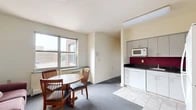 Located at 327 Eddy Street in Ithaca, NY, this 3-bedroom, 1-bathroom unit offers a practical and well-organized single-floor layout. The kitchen is a standout feature, equipped with gray cabinetry extending to the upper walls, dark granite-style countertops, a bold crimson tile backsplash, and white appliances including a refrigerator, electric range, dishwasher, and microwave. Light tile flooring completes the kitchen's clean, functional aesthetic. An adjacent dining area with a round wooden table and chairs sits beside a large window, allowing natural light to fill the space. Each of the three carpeted bedrooms features large windows that frame views of the Ithaca skyline and surrounding greenery. The bedrooms include built-in closets and are furnished with beds, dressers, desks, and shelving units, providing ample storage. The shared full bathroom includes a tub-shower combination with sliding glass doors, a vanity with under-sink storage, and light tile flooring. A hallway connects all rooms, contributing to a straightforward and efficient floor plan. The unit's elevated position provides pleasant outdoor views, adding to the overall appeal of this Ithaca residence.