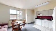 Located at 327 Eddy Street in Ithaca, NY, this 3-bedroom, 1-bathroom unit offers a practical and well-organized single-floor layout. The kitchen is a standout feature, equipped with gray cabinetry extending to the upper walls, dark granite-style countertops, a bold crimson tile backsplash, and white appliances including a refrigerator, electric range, dishwasher, and microwave. Light tile flooring completes the kitchen's clean, functional aesthetic. An adjacent dining area with a round wooden table and chairs sits beside a large window, allowing natural light to fill the space. Each of the three carpeted bedrooms features large windows that frame views of the Ithaca skyline and surrounding greenery. The bedrooms include built-in closets and are furnished with beds, dressers, desks, and shelving units, providing ample storage. The shared full bathroom includes a tub-shower combination with sliding glass doors, a vanity with under-sink storage, and light tile flooring. A hallway connects all rooms, contributing to a straightforward and efficient floor plan. The unit's elevated position provides pleasant outdoor views, adding to the overall appeal of this Ithaca residence.