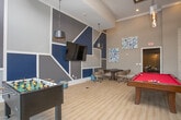 This thoughtfully designed game and entertainment lounge offers an engaging, multi-functional space ideal for socializing and recreation. The room's most striking architectural feature is its soaring ceiling height, which lends an open, expansive feel and allows natural light to pour in through tall windows, brightening the light wood-look flooring that runs throughout.

At the center of the space sits a full-size billiards table with a bold red felt surface, serving as the room's focal point. Nearby, an arcade gaming console and a foosball table provide additional entertainment options. A wall-mounted flat-screen television is positioned alongside built-in shelving accented in navy blue, adding both style and functionality.

A dedicated lounge area features a sleek, linear fireplace framed by a shimmering mosaic tile surround, with a wall-mounted television above it. Contemporary seating, including a sculptural black swivel chair and tan leather ottomans, creates a cozy retreat within the larger space. Exposed wooden columns add warmth and contrast against the cool gray and navy palette.

A round table with rolling chairs offers a flexible area for casual gatherings or tabletop games. Pendant lighting fixtures in industrial-style finishes complete the modern, curated aesthetic of this well-appointed entertainment space.