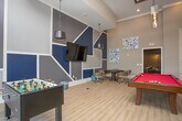 This thoughtfully designed game and entertainment lounge offers an engaging, multi-functional space ideal for socializing and recreation. The room's most striking architectural feature is its soaring ceiling height, which lends an open, expansive feel and allows natural light to pour in through tall windows, brightening the light wood-look flooring that runs throughout.

At the center of the space sits a full-size billiards table with a bold red felt surface, serving as the room's focal point. Nearby, an arcade gaming console and a foosball table provide additional entertainment options. A wall-mounted flat-screen television is positioned alongside built-in shelving accented in navy blue, adding both style and functionality.

A dedicated lounge area features a sleek, linear fireplace framed by a shimmering mosaic tile surround, with a wall-mounted television above it. Contemporary seating, including a sculptural black swivel chair and tan leather ottomans, creates a cozy retreat within the larger space. Exposed wooden columns add warmth and contrast against the cool gray and navy palette.

A round table with rolling chairs offers a flexible area for casual gatherings or tabletop games. Pendant lighting fixtures in industrial-style finishes complete the modern, curated aesthetic of this well-appointed entertainment space.