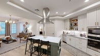 Located at 98 South River Drive in Tempe, Arizona, this thoughtfully designed 2-bedroom, 2-bathroom residence offers a cohesive blend of modern finishes and functional living across a single floor. The kitchen serves as a standout centerpiece, featuring soft sage-green cabinetry accented with brushed gold hardware, granite countertops, a white subway tile backsplash, stainless steel appliances, and a large island with seating — all complemented by a striking stainless steel range hood overhead. Light wood-look flooring flows throughout, tying the spaces together seamlessly. The living room offers natural light through dual windows, enhanced by a statement brass chandelier and tray ceiling detailing. The primary bedroom is generously sized, offering a ceiling fan and window, while its walk-in closet provides dual hanging rods and built-in shelving for ample storage. The second bedroom is equally comfortable with its own closet. Both bathrooms feature granite vanity tops, deep green cabinetry with gold hardware, subway tile surrounds, and soaking tub-shower combinations. Each closet is well-organized with built-in shelving systems. The overall ambiance is modern yet warm, with refined design details carried consistently throughout the home.