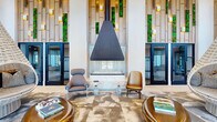 This expansive lobby in Annapolis, Maryland, spans a generous single floor and delivers a striking first impression through its sophisticated, contemporary design. The entrance area is anchored by a dramatic floor-to-ceiling feature wall clad in warm wood paneling, accented by vertical strips of vibrant living moss and gold wall sconces that create a natural yet refined aesthetic. A large, statement suspended fireplace with a marble surround serves as a central focal point, flanked by designer lounge chairs and glass-and-crystal accent tables atop an abstract area rug.

The main corridor extends deep into the space, drawing the eye through a series of curated seating zones. Oversized suspended wicker pod chairs hang from the soaring ceiling, providing intimate, cocoon-like seating beneath clusters of globe pendant lights. Light-toned large-format tile flooring runs throughout, reinforcing the bright, airy atmosphere. Built-in window seat alcoves framed by wood millwork offer views to the outdoors, complemented by modern upholstered chairs and side tables.

Toward the rear, a separate lounge and kitchenette area features soaring angled ceilings, an ornate gold mosaic wall panel, and casual dining seating beneath sculptural pendant lighting, completing the lobby's layered, resort-inspired ambiance.