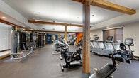 This fitness center offers a comprehensive workout environment spread across a single, open floor. The space is defined by its striking exposed wood beam columns and posts, which add a warm, structural character to the otherwise modern interior. Bold accent walls in deep blue and vibrant orange energize the space and help delineate distinct workout zones.

The cardio area is generously stocked with a full row of treadmills, elliptical machines, stationary bikes, stair climbers, and cycling bikes, providing ample options for aerobic training. Adjacent to this zone, a dedicated strength training area houses a multi-station cable machine and a variety of resistance equipment. A separate functional training alcove features TRX suspension trainers, medicine balls, kettlebells, a BOSU ball, resistance bands, and a weight bench, with floor-to-ceiling mirrors enhancing the sense of space and allowing for form checks during workouts.

The flooring throughout is durable rubber gym matting, practical and suited for high-intensity use. Overhead, bright recessed and linear lighting ensures excellent visibility across all areas. A set of glass doors opens directly to an outdoor pool and lounge area, offering natural light and a seamless indoor-outdoor connection. The overall ambiance is professional, energetic, and thoughtfully organized to support a wide range of fitness activities.