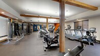 This fitness center offers a comprehensive workout environment spread across a single, open floor. The space is defined by its striking exposed wood beam columns and posts, which add a warm, structural character to the otherwise modern interior. Bold accent walls in deep blue and vibrant orange energize the space and help delineate distinct workout zones.

The cardio area is generously stocked with a full row of treadmills, elliptical machines, stationary bikes, stair climbers, and cycling bikes, providing ample options for aerobic training. Adjacent to this zone, a dedicated strength training area houses a multi-station cable machine and a variety of resistance equipment. A separate functional training alcove features TRX suspension trainers, medicine balls, kettlebells, a BOSU ball, resistance bands, and a weight bench, with floor-to-ceiling mirrors enhancing the sense of space and allowing for form checks during workouts.

The flooring throughout is durable rubber gym matting, practical and suited for high-intensity use. Overhead, bright recessed and linear lighting ensures excellent visibility across all areas. A set of glass doors opens directly to an outdoor pool and lounge area, offering natural light and a seamless indoor-outdoor connection. The overall ambiance is professional, energetic, and thoughtfully organized to support a wide range of fitness activities.