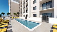 This property is part of a contemporary multi-story building characterized by a crisp white exterior facade accented with black-railed balconies and arched ground-level architectural details. The outdoor amenity area is a standout feature, centered around a generously sized lap pool finished with blue tile and framed by large-format light stone pavers that extend throughout the surrounding deck. Yellow lounge chairs are positioned poolside, offering a vibrant contrast to the neutral tones of the building exterior and hardscape. Tropical palm trees are visible at the far end of the pool area, adding a resort-like ambiance appropriate to the warm-climate setting. The pool area is flanked on multiple sides by the building's facade, creating an enclosed, private courtyard atmosphere. Gated access points are visible, enhancing both security and definition of the outdoor space. The building rises several stories above the amenity level, with each floor featuring private balconies. The overall design aesthetic is clean, modern, and functional, with an emphasis on outdoor living and communal amenities. Interior images were not sufficiently illuminated to provide additional details.