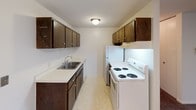 This single-floor unit offers a functional and straightforward layout across several well-defined rooms. The living area is generously sized, featuring wall-to-wall carpeting in a warm brown tone and clean white walls that lend a bright, neutral backdrop. Baseboard heating units run along the perimeter walls, and vertical blinds cover a sliding glass door that brings in natural light. A ceiling fan in an adjacent room provides additional air circulation and lighting.

The galley-style kitchen is efficiently arranged, with dark wood cabinetry lining both sides of the space, offering ample upper and lower storage. White appliances, including a coil-burner electric range and a refrigerator, are paired with a double-basin stainless steel sink set into a light-toned countertop. A flush ceiling light illuminates the space, and the floor is finished in a neutral vinyl tile.

The bathroom presents a clean, classic design with white square wall tiles surrounding a full-size soaking tub and shower combination. The floor is finished in white tile, and the room includes a toilet, vanity with countertop, and towel bars. A natural wood-finish door adds warmth to the entry.

The bedrooms are carpeted and bright, with windows fitted with horizontal blinds offering outdoor views and natural light, complemented by baseboard heaters beneath each window.
