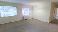 This single-floor space offers a clean, understated aesthetic defined by neutral tones and thoughtful design details. The main living area is generously proportioned, featuring wall-to-wall plush carpeting in a warm beige tone that adds comfort underfoot. Soft, cream-colored walls create a bright and airy atmosphere throughout the room.

Natural light is a standout feature, with multiple windows allowing sunlight to fill the space and offer views of the surrounding greenery outside. A baseboard heating unit is positioned beneath the central window, ensuring year-round comfort. An elegant chandelier with a classic multi-arm design serves as a focal point overhead, lending a touch of refinement to the otherwise simple interior.

The layout is open and flexible, providing ample room to arrange furniture in a variety of configurations to suit different needs. Clean white baseboards frame the carpeted floors, adding a crisp finishing detail to the room. The overall ambiance is calm and inviting — a well-maintained, move-in-ready space that balances simplicity with functionality, making it suitable for a range of uses and lifestyles.