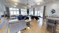 This single-floor community clubroom is designed for both relaxation and entertainment, offering an inviting and well-appointed gathering space. The room features light hardwood flooring throughout, which complements the neutral-toned walls and tray ceiling details. Large windows and glass doors line multiple walls, bathing the interior in abundant natural light while providing views of the surrounding outdoor areas. Ceiling fans are strategically placed to ensure comfort throughout the space.

The lounge area is anchored by plush seating, including a bold navy blue sectional sofa paired with a lighter gray sofa and a central coffee table, creating a comfortable conversation zone. A stylish area rug adds warmth and definition to the seating arrangement. Adjacent to the lounge is a striking dark-finished pool table, serving as a central recreational focal point of the room. Bar-height seating and additional casual furniture are positioned nearby, offering flexible options for gathering.

Decorative drum pendant lighting and draping gray curtains add a polished, contemporary aesthetic. The open layout promotes easy movement and accommodates a variety of social activities. Overall, the space exudes a modern, welcoming ambiance well-suited for community events, casual hangouts, and recreational use.
