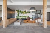 This thoughtfully designed single-floor space offers a generous, open-plan layout encompassing a living room, dining room, and kitchen. The interior showcases a refined modern aesthetic, blending natural wood accents with sleek tile, concrete, and glass elements throughout.

The entry foyer makes an immediate impression with floor-to-ceiling glass curtain walls that flood the space with natural light and frame views of the surrounding landscape. Woven leather benches and a potted fiddle-leaf fig tree add warmth to the minimalist entryway.

The living area features large-format tile flooring, a natural wood structural column, and a lush vertical garden installation. A neutral sectional sofa and concrete coffee table anchor the lounge zone, complemented by pendant greenery above. Steel-framed glass partition walls define adjacent rooms while preserving an open, airy feel.

The dining area is highlighted by a solid wood communal table, modern seating, and a striking slatted wood ceiling detail with exposed ductwork that lends an industrial-chic character. Large black-framed windows offer tree-lined outdoor views, filling the room with natural light.

The kitchen zone features sleek navy cabinetry with white countertops, set against a dramatic floor-to-ceiling textured tile accent wall with integrated sconce lighting. Throughout, the cohesive material palette of wood, tile, and concrete creates a warm yet contemporary ambiance suited for both collaboration and relaxation.