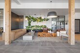 This thoughtfully designed single-floor space offers a generous, open-plan layout encompassing a living room, dining room, and kitchen. The interior showcases a refined modern aesthetic, blending natural wood accents with sleek tile, concrete, and glass elements throughout.

The entry foyer makes an immediate impression with floor-to-ceiling glass curtain walls that flood the space with natural light and frame views of the surrounding landscape. Woven leather benches and a potted fiddle-leaf fig tree add warmth to the minimalist entryway.

The living area features large-format tile flooring, a natural wood structural column, and a lush vertical garden installation. A neutral sectional sofa and concrete coffee table anchor the lounge zone, complemented by pendant greenery above. Steel-framed glass partition walls define adjacent rooms while preserving an open, airy feel.

The dining area is highlighted by a solid wood communal table, modern seating, and a striking slatted wood ceiling detail with exposed ductwork that lends an industrial-chic character. Large black-framed windows offer tree-lined outdoor views, filling the room with natural light.

The kitchen zone features sleek navy cabinetry with white countertops, set against a dramatic floor-to-ceiling textured tile accent wall with integrated sconce lighting. Throughout, the cohesive material palette of wood, tile, and concrete creates a warm yet contemporary ambiance suited for both collaboration and relaxation.