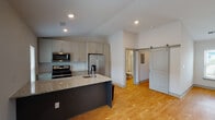 Located in Huntsville, AL (35806), this single-floor space offers a thoughtfully designed interior with modern finishes throughout. The kitchen serves as a central highlight, featuring a large island with granite countertops, a deep sink, and a full suite of stainless steel appliances including a gas range, built-in microwave, French door refrigerator with ice maker, and dishwasher. Crisp white upper cabinetry pairs with darker lower cabinets for a contemporary contrast, while recessed lighting and vaulted ceiling details add a sense of openness. Warm-toned hardwood flooring flows seamlessly throughout the main living areas, complementing the bright, neutral wall palette. A stylish sliding barn door adds a distinctive architectural touch, providing access to the bathroom. The bathroom is clean and modern, equipped with a granite-topped vanity, framed mirror, walk-in glass-enclosed shower, and wood-toned flooring. The open living area benefits from natural light through multiple windows with blinds. A carpeted stairwell with dual handrails provides access to additional levels. Overall, the space presents a clean, contemporary aesthetic with quality finishes and functional design.