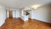 This contemporary single-floor residence offers a thoughtfully designed layout with a clean, modern aesthetic throughout. The open-concept living and dining area is anchored by rich hardwood flooring and floor-to-ceiling sliding glass doors that lead to a balcony, flooding the space with natural light and providing sweeping panoramic views of the surrounding cityscape. Dark curtain panels frame the expansive windows, adding a refined contrast to the bright, white walls and ceiling.

The kitchen is designed in a galley configuration, featuring gray flat-front cabinetry lining both walls, light granite-style countertops, a dark tile backsplash, and stainless steel appliances including a refrigerator, electric range, and built-in microwave. A peninsula counter connects the kitchen to the living area, providing additional workspace and serving as a natural divider between the two zones.

The bedrooms are carpeted in a neutral tone and offer generous closet space with built-in rods. The primary bedroom also benefits from floor-to-ceiling glass doors opening to the balcony, further enhancing the airy, elevated feel of the space. The bathroom is clean and functional, finished in white tile with a full bathtub and shower combination. Throughout the unit, the consistent use of white walls, warm wood flooring, and modern fixtures creates a cohesive and inviting atmosphere.