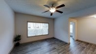 This single-floor space offers a straightforward and functional layout designed for comfortable everyday living. The main living area is a bright, open room finished with durable wood-look plank flooring in a rich gray-toned finish that runs throughout the space, lending a clean and cohesive aesthetic. A large picture window, fitted with horizontal blinds, anchors one wall and draws in generous amounts of natural light, creating an airy and welcoming atmosphere. Overhead, a classic five-blade ceiling fan with a built-in light fixture provides both ventilation and ambient lighting. The walls are finished in a neutral tone, complementing the textured popcorn ceiling and providing a versatile backdrop for a variety of décor styles. An open archway connects the main living area to an adjacent room, also featuring matching flooring and a window, suggesting a seamless and well-connected floor plan. The overall design is practical and understated, making the space adaptable to a range of uses. The consistent flooring, neutral color palette, and natural light throughout contribute to a bright, inviting, and move-in-ready environment.