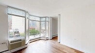 Located at 55 West Chestnut Street in Chicago's Near North Side, this one-bedroom, one-bathroom residence offers a well-organized layout across a single floor. The living room is a standout space, highlighted by a striking curved bay window that wraps the corner and floods the room with natural light while framing views of the surrounding Chicago cityscape and greenery. Light hardwood flooring runs throughout the main living areas and bedroom, lending warmth and continuity to the interior.

The bedroom is a comfortable, well-proportioned room featuring its own ceiling fan, baseboard heating, and access to a dedicated walk-in closet equipped with wire shelving for organized storage. A tiled hallway connects the living areas to the kitchen and bathroom, providing a clean transition between spaces. The kitchen includes white cabinetry, dark countertops, and full-sized appliances including a refrigerator. The bathroom is finished with large-format tile surrounding a full soaking tub and shower combination, with a mirrored medicine cabinet above the vanity. Clean white walls and a bright, neutral palette throughout contribute to an open and airy ambiance well-suited to urban living.