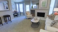 Spanning three floors, this residence offers a thoughtfully designed layout with distinct living and dining areas on the main level. The living room centers around a white-tiled fireplace, which serves as a focal point and adds a classic, cozy element to the space. Decorative vases and greenery adorn the mantle, complementing the neutral, understated aesthetic. A pair of modern wire-base coffee tables provide both function and visual interest against the plush, wall-to-wall carpeting. A cushioned armchair with turned legs adds a traditional touch near the hearth, while woven storage baskets contribute to an organized, tidy atmosphere. Natural light filters generously into the space through large bay windows dressed in bold chevron-patterned drapes, creating a bright and airy feel. The adjacent dining area, visible from the living room, features dark-framed chairs and a coordinating table, offering a seamless flow between entertaining spaces. Soft, neutral wall tones throughout lend a calm, cohesive ambiance, making the main level feel both welcoming and well-appointed.