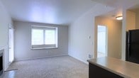 This single-floor residence presents a straightforward and functional layout designed for comfortable everyday living. The main living area is finished with wall-to-wall carpet in a neutral beige tone, creating a warm and understated atmosphere throughout the space. Smooth, light-colored walls pair with white trim and baseboards to give the interior a clean, cohesive look. A centrally positioned window allows natural light to filter into the living area, brightening the space and offering a view of the greenery outside. A floor vent indicates a forced-air climate control system, ensuring year-round comfort. The living area flows naturally toward an adjacent room, visible through a doorway that hints at additional carpeted space beyond — suggesting a bedroom or secondary living area. A partial view of a dark-toned countertop in the foreground suggests proximity to a kitchen or bar area, adding to the functional versatility of the floor plan. The overall ambiance is quiet and neutral, providing a blank canvas well-suited for a variety of interior styles. With its simple finishes, connected layout, and ample natural light, this space offers a practical and livable environment.