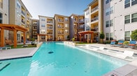 This expansive outdoor amenity space is the centerpiece of a contemporary multi-story residential community, offering a resort-inspired environment designed for relaxation and recreation. The focal point is a generously sized swimming pool with crystal-clear blue water, complemented by an adjoining spa and integrated in-pool sun ledges with lounge chairs — ideal for unwinding in the sun. The pool deck is finished in smooth concrete and is surrounded by a thoughtfully landscaped courtyard that includes manicured greenery, ornamental trees, and decorative stone accents. A striking wooden pergola serves as an architectural focal point, providing a shaded gathering area adjacent to the pool. Multiple lounge chairs, seating areas, and umbrellas are arranged throughout the deck to accommodate residents at every preference. The surrounding multi-story building features a U-shaped design with private balconies on each floor overlooking the courtyard, creating an intimate and enclosed atmosphere. Floor-to-ceiling windows on the ground level allow natural light to flow into interior amenity spaces while maintaining visual connectivity with the outdoor area. A secondary courtyard with additional seating and landscaping offers a quieter retreat. Together, these elements create an inviting, community-oriented outdoor living space that balances aesthetics with functionality.