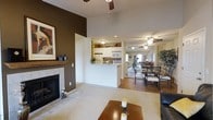 This single-floor space showcases an open-concept layout that seamlessly connects the living room, kitchen, and dining area into one cohesive environment. The living room serves as a focal point, anchored by a tiled fireplace framed by a classic wood mantle and set against a rich, dark brown accent wall. A ceiling fan overhead and plush carpeted flooring contribute to the room's comfortable, inviting character. A wooden coffee table with candle accents sits at the center, complementing the warm tones throughout.

The kitchen features a pass-through design with white cabinetry extending to the upper walls, a built-in microwave, and white appliances. Open shelving above the pass-through adds a display element, while the layout maximizes efficiency in a compact footprint.

Adjacent to the kitchen, the dining area transitions to hardwood flooring and accommodates a full dining set with dark metal chairs. A ceiling fan provides air circulation, and a window at the far end draws in natural light, brightening the space. The overall ambiance is warm and casual, with a cohesive neutral and earth-toned palette tying all areas together.