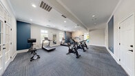 This fitness center is organized across two connected rooms, offering a comprehensive workout environment designed for a variety of exercise routines. The main exercise area is generously proportioned, featuring plush, blue-toned carpet tiles that provide a comfortable and durable surface underfoot. Multiple spinning bikes and treadmills are arranged throughout the space, complemented by a rowing machine and a wall-mounted television for entertainment during cardio sessions. Large windows along the perimeter flood the room with natural light, while recessed ceiling lights and exposed beam detailing add a polished architectural touch. A full-length mirror reflects the open layout, enhancing the sense of space. Distinctive navy blue accent walls provide a bold contrast to the otherwise neutral, light gray palette throughout. French doors connect the main cardio room to a secondary functional training room, which offers a quieter, more focused setting. This adjacent room features additional natural light from double windows, crown molding, and transom window detailing above the doorway. It is equipped with a plyo box and a rack stocked with stability balls and resistance bands, making it ideal for stretching, strength training, and functional fitness exercises. Together, the two rooms create a versatile and well-appointed fitness facility.