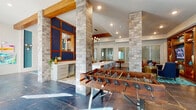 Located at 646 W Smith St in Orlando, FL, this single-floor community amenity space offers a dynamic and thoughtfully curated environment designed for socializing and recreation. The open layout is defined by striking exposed stone columns, warm wood beams, and a mix of slate tile and hexagonal stone flooring that establish a distinct architectural character throughout.

The game area takes center stage with a classic foosball table, complemented by a large-format Connect Four game nearby, making it an inviting hub for interactive entertainment. Bold, large-scale swan paintings in vivid teal tones adorn the whitewashed brick walls, adding an artistic focal point to the lounge seating area, which features a curved teal sofa and a glass-top coffee table.

A cozy media lounge is anchored by a built-in wood entertainment wall with integrated shelving, accent lighting, a mounted television, and plush navy ottomans set atop a vibrant teal geometric rug. Adjacent to the game area, a well-appointed kitchenette features a white quartz island with stylish bar seating, modern cabinetry with brass hardware, an under-counter beverage refrigerator, and a bold navy-and-wood paneled accent wall. Pendant globe lighting adds warmth throughout. Natural light flows in generously through large windows and glass doors, enhancing the space's energetic yet welcoming ambiance.