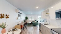 This thoughtfully designed single-floor studio offers a clean, contemporary aesthetic throughout. The open-concept layout seamlessly connects the kitchen, dining, living, and sleeping areas, creating an efficient and cohesive living environment bathed in natural light from multiple windows and a sliding glass door.

The kitchen is well-appointed with white upper cabinetry, light wood-toned lower cabinets, white quartz countertops, a stainless steel sink, electric range, built-in microwave, and a full-size refrigerator — providing a complete set of appliances for everyday cooking.

The main living area flows naturally into the sleeping space, with light-toned hardwood-style flooring extending throughout. A dedicated workspace with a mid-century modern desk adds functional versatility. The sleeping area accommodates a full-size bed alongside a green tufted sofa, and features a sliding glass door opening directly to a private outdoor patio.

The bathroom is clean and minimal, finished with light flooring and a shower/tub combination. A wall-mounted mini-split HVAC unit provides climate control. Recessed lighting and crisp white walls contribute to a bright, airy ambiance throughout the entire unit.