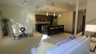 This two-story space showcases a thoughtfully designed open-concept main living area that seamlessly integrates the kitchen, dining, and living zones into one cohesive environment. The kitchen is outfitted with dark espresso cabinetry, stainless steel appliances including a refrigerator and microwave, and dark granite countertops. A central island with bar-height seating accommodates several stools, making it a functional and social focal point. Pendant lighting in cylindrical white shades descends at varying heights above the island, adding a refined decorative touch. Recessed lighting and track lighting complement the overall illumination throughout the space. The dining area features a glass-top table paired with clean-lined white chairs, positioned adjacent to the kitchen for convenient entertaining. The living area, visible in the foreground, is furnished with a generously sized white sofa accented with patterned throw pillows. Light-toned carpeting flows throughout, contrasting effectively with the dark kitchen finishes. The layout promotes an open, airy feel, with the upper floor offering additional living space. Overall, the design balances contemporary aesthetics with practical functionality.
