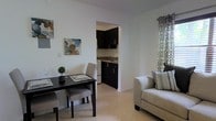 This single-floor space offers a thoughtfully arranged layout that maximizes functionality and comfort. The living area is bright and welcoming, featuring crisp white walls that amplify the natural light filtering through patterned curtains. Light-toned ceramic tile flooring runs throughout, providing a clean and cohesive look across the entire unit.

The dining area is positioned adjacent to the living space, comfortably accommodating a dark-toned dining table with upholstered chairs — an ideal spot for everyday meals. A small decorative plant adds a touch of greenery to the dining setup, complemented by colorful wall art that brings personality to the otherwise neutral palette.

The kitchen is accessible through an arched passageway, giving the space an open yet defined sense of separation. It features dark cabinetry with contrasting countertops, offering ample storage and prep space. White appliances complete the kitchen, contributing to a clean and practical cooking environment.

The living area includes a light gray sofa accented with patterned throw pillows, creating a relaxed and inviting atmosphere. The overall design balances simplicity with style, using a neutral color palette with deliberate accents to create a space that feels both modern and comfortable.