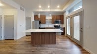 This thoughtfully designed single-floor residence combines contemporary style with functional living spaces. The open-concept layout connects the kitchen and living area seamlessly, with warm wood-look flooring flowing throughout the main areas, complemented by a ceiling fan with integrated lighting.

The kitchen is a standout feature, showcasing rich walnut-toned cabinetry with sleek hardware, a white quartz countertop, and a large center island with pendant lighting above. A classic white subway tile backsplash frames the stainless steel appliances, including a refrigerator, range, microwave, and dishwasher. Natural light pours in through a full-length glass door and transom windows.

The bedrooms offer a quieter retreat, featuring plush wall-to-wall carpeting in one room and wood-look flooring in another, each with windows dressed in horizontal blinds that allow for adjustable natural light. The bathrooms maintain the home's cohesive aesthetic with matching walnut vanity cabinetry, white quartz countertops, large frameless mirrors, and contemporary chrome fixtures. The flooring transitions to wood-look planks, adding warmth and durability. Overall, the space presents a clean, modern ambiance with consistent, high-quality finishes throughout.