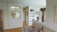 This single-floor residence offers a thoughtfully arranged open-concept layout that connects the living, dining, and kitchen areas into one cohesive and functional space. The living area is anchored by warm hardwood flooring that extends throughout, providing a welcoming and consistent aesthetic underfoot. A standout feature is the built-in white shelving unit that serves as both a display and organizational focal point, offering tiered shelves ideal for décor, books, and personal accents.

The kitchen is accessible via a raised pass-through counter with an arched surround, creating a semi-open connection between the cooking and living spaces. This design element enhances both visibility and interaction while maintaining a subtle sense of separation. The kitchen itself features white appliances and ample counter space, keeping the look clean and practical.

Neutral-toned walls in soft beige and sage green tones contribute to a calm and inviting atmosphere throughout. Arched doorways lead to additional rooms, adding an architectural detail that lends character to the overall design. The ceilings are fitted with flush-mount lighting, maintaining a clean and uncluttered overhead profile. The overall ambiance is warm, functional, and well-suited for everyday living.
