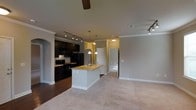 Located at 2015 Copperleaf Pkwy in Durham, North Carolina, this single-floor residence offers a thoughtfully designed open-concept layout that seamlessly connects the kitchen and living areas. The kitchen is a standout feature, anchored by a central island with a light stone countertop and an undermount sink, providing both workspace and a casual gathering spot. Dark cabinetry with sleek hardware creates a bold contrast against the lighter countertops and walls, while a suite of black appliances — including a refrigerator, range, and built-in microwave — adds a cohesive, contemporary feel. Track lighting and pendant lights overhead ensure the kitchen is well-lit and functional. Crown molding runs throughout the space, lending an elegant, polished touch to the interiors. The living area features plush carpeting in a warm neutral tone, offering a comfortable transition from the hardwood-style flooring in the kitchen. A ceiling fan is positioned centrally in the living space, adding to the comfort of the home. Arched architectural details frame doorways, adding character and visual interest. A hallway leads to additional rooms, including what appears to be a well-appointed bathroom, rounding out this inviting and practical single-floor home.
