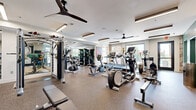 Located at 19501 W 102nd St in Lenexa, Kansas, this well-appointed fitness center offers a generously sized, single-floor exercise facility designed to accommodate a full range of workout routines. The space features durable speckled rubber flooring throughout, providing both safety and comfort during exercise. Large windows along the perimeter flood the room with natural light and offer views of the surrounding outdoor environment, creating an energizing atmosphere for workouts.

The main exercise area is comprehensively equipped with cardio machines including treadmills, ellipticals, stationary bikes, and a recumbent bike. A dedicated strength training section houses a multi-station cable machine, weight benches, and a full dumbbell rack, catering to a variety of resistance training needs. Ceiling fans and recessed linear lighting ensure a comfortable and well-lit environment throughout.

A standout feature is the glass-enclosed spin studio, which offers a private space for cycling workouts on dedicated spin bikes, with wood-look flooring and teal accent walls adding a stylish touch. The main room also features teal accent walls, motivational artwork including a neon boxing-themed piece, and a wall-mounted television. Multiple ceiling fans maintain airflow, and the open layout ensures ample space for movement between equipment stations.