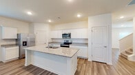 Located at 5107 Coopers Landing Rd in Summerville, South Carolina, this two-story home offers a clean, contemporary design with thoughtful finishes throughout. The main level showcases wood-look plank flooring that flows seamlessly through the open living areas and into the well-appointed kitchen. The kitchen is a standout feature, centered around a large island with granite countertops and a built-in sink. White shaker-style cabinetry lines the walls, complemented by stainless steel appliances including a refrigerator, electric range, and over-the-range microwave. Recessed lighting illuminates the space, enhancing its bright, open feel. The upper level transitions to plush carpeting in the bedrooms, each benefiting from natural light through window blinds. A spacious walk-in closet with wire shelving provides generous storage. The upper hallway connects the rooms with ease, featuring the same wood-look flooring and staircase railing detail seen on the main floor. A full bathroom on the upper level includes a tub-shower combination with tile surround, wood-look tile flooring, and a white vanity. Throughout the home, neutral tones and recessed lighting create a consistent, move-in-ready ambiance suited for comfortable everyday living.