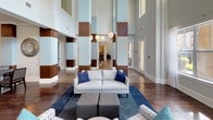 This single-floor community space is designed with an open, expansive layout that makes a strong architectural statement from the moment you enter. Soaring double-height ceilings define the main lounge area, creating an airy, grand atmosphere accentuated by an abundance of natural light streaming through large multi-pane windows. Structural columns clad in warm wood paneling and accented with soft blue hues add a distinctive design element that ties the color palette together throughout the space.

Rich, dark hardwood flooring extends seamlessly across the entire floor, grounding the space with a polished, upscale feel. The primary lounge area is furnished with a contemporary light gray sofa, patterned accent chairs, and a deep navy area rug, offering a comfortable and inviting setting for socializing or relaxing. Pendant globe lighting adds a warm, modern touch overhead.

A dedicated recreation area includes a full-size pool table and a large wall-mounted flat-screen television, creating an ideal entertainment zone. Adjacent to this, a café-style seating nook features a round bistro table and upholstered chairs beneath recessed lighting and elegant crown molding. Curated abstract artwork adorns the walls, adding a refined, gallery-like quality. French doors open directly to lush outdoor greenery, further enhancing the connection between interior comfort and the natural surroundings.