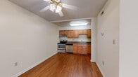This single-floor residence offers a straightforward and functional layout connecting several distinct living areas. The main living space is anchored by warm hardwood flooring and neutral white walls, creating a bright and open atmosphere. A ceiling fan with a light fixture overhead provides both air circulation and ambient lighting. One wall features exposed brick detailing near the entryway, adding a subtle textural accent to the otherwise clean interior.

The kitchen is positioned at one end of the living area and is equipped with warm-toned wood cabinetry offering ample upper and lower storage, dark granite-style countertops, and stainless steel appliances including an electric range and dishwasher. A refrigerator and under-cabinet lighting round out the kitchen's practical features. Light tile flooring and a flush-mounted ceiling fixture illuminate the workspace effectively.

A carpeted hallway connects the living area to the bedroom, which features plush neutral carpeting, a ceiling fan, and a window allowing natural light. A louvered closet door provides accessible storage. The bathroom is tiled in light tones and includes a walk-in shower with white tile surround and a standard toilet, maintaining a clean and functional design throughout.