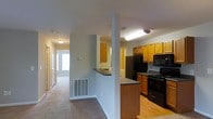 This single-floor residence offers a practical and well-organized layout that connects the main living areas in a seamless flow. The kitchen is a standout feature, equipped with warm honey-toned wood cabinetry that provides ample storage, a full suite of black appliances including a refrigerator, electric range, and overhead microwave, and a peninsula-style counter that serves as both a workspace and a casual dining surface. The hard-surface flooring in the kitchen transitions smoothly into carpeted living areas, creating a clear distinction between functional and relaxation zones. A structural column adds architectural character to the open transition space between the kitchen and the hallway leading to additional rooms. The hallway connects the living spaces to what appears to be a well-lit bedroom or secondary room, visible at the far end with natural light filtering through a window. The neutral color palette of soft grays and warm wood tones throughout gives the space a clean, cohesive appearance. Overhead lighting is present throughout, ensuring consistent illumination across all areas. A floor vent is prominently placed along the baseboard in the main living corridor, reflecting a centralized HVAC system. Overall, the space presents a straightforward, functional design suited for comfortable daily living.