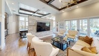 Located at 480 John Carter Rd in Savannah, GA, this thoughtfully designed single-floor community space offers an inviting open-concept layout that seamlessly blends comfort and functionality. The soaring coffered ceilings with exposed dark wood beams create a striking architectural focal point, complemented by warm hardwood flooring throughout. A dramatic shiplap accent wall anchors the living area, which features a large mounted flat-screen TV and a comfortable seating arrangement of tufted sofas and modern accent chairs arranged around a central coffee table. Natural light pours in through an expansive wall of floor-to-ceiling windows, offering pleasant views of the surrounding greenery and neighborhood. The adjacent game area includes a full-size billiards table positioned beneath abundant windows that frame outdoor views. The kitchen is a standout feature, showcasing gray cabinetry, white quartz countertops, a stainless steel refrigerator, and a vibrant teal ceramic tile backsplash that adds a bold design element. A dedicated dining area offers restaurant-style booth seating with reclaimed wood tables and modern pendant lighting, creating an intimate yet functional space for gatherings. Together, these spaces deliver a cohesive, upscale aesthetic with a warm, welcoming ambiance.