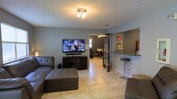 This single-floor space is designed with an open-concept layout that seamlessly connects the living area to a kitchen bar counter, creating a functional and social atmosphere. The living room is finished with light ceramic tile flooring and grey-painted walls, offering a neutral and versatile backdrop. A textured ceiling with a central flush-mount light fixture provides even illumination throughout the room. The living area is spacious enough to comfortably accommodate a sectional sofa and ottoman, positioned to face a mounted flat-screen television set atop a media console. Adjacent to the living room, a raised breakfast bar with granite-style countertop and bar stools provides a casual dining and gathering spot, separated from the kitchen by a partial wall with a pass-through opening. A large mirror is mounted above the bar area, adding depth and visual openness to the space. A doorway visible from the living room leads to an additional area, which features its own ceiling fixture and tiled flooring, suggesting a connected room or hallway. The overall ambiance is casual and practical, with a cohesive grey-and-neutral color palette tying the entire single-floor layout together.