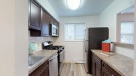 This single-floor unit is thoughtfully laid out across several distinct areas. The galley-style kitchen is equipped with stainless steel appliances, including a gas range with overhead microwave, a full-size refrigerator, and a stainless sink. Dark espresso cabinetry provides ample storage, complemented by granite-toned countertops and a classic subway tile backsplash. Light wood-style flooring runs throughout the kitchen, and a window with blinds allows natural light to brighten the space. A pass-through opening connects the kitchen to the main living area.

The living area is generously sized with neutral wall-to-wall carpeting, a ceiling fan with light fixture, and large windows that invite natural light. A wall-mounted air conditioning unit and baseboard heating ensure year-round comfort. The layout connects seamlessly to additional rooms, including what appears to be a spacious bedroom also featuring carpet flooring, a ceiling fan, a window with blinds, baseboard heating, and a wall-mounted AC unit.

The bathroom is clean and functional, featuring a full-size soaking tub with a tiled shower surround and chrome fixtures. Overall, the unit presents a practical, well-maintained interior with consistent neutral tones throughout.